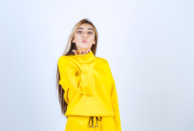 Young woman in yellow sweatsuit blowing kiss over white wall