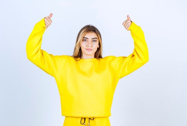 Young woman in yellow sweatsuit giving thumbs up on white wall