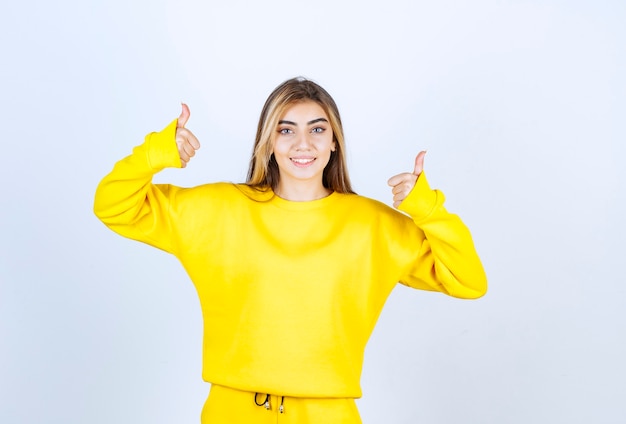 Young woman in yellow sweatsuit giving thumbs up on white wall