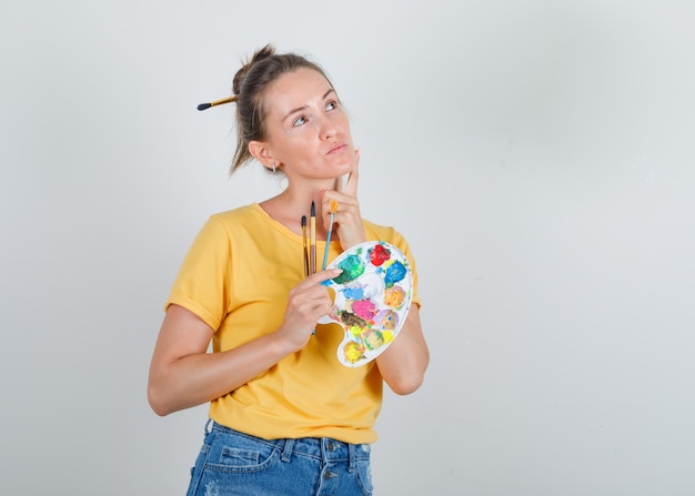 Young woman in yellow t-shirt, jeans looking up and holding painting tools