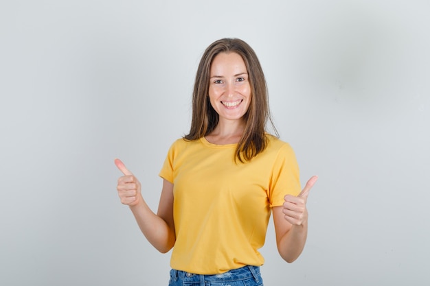 Young woman in yellow t-shirt, shorts showing thumbs up and looking merry