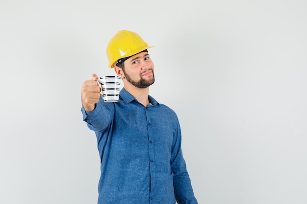 Young worker offering a cup of coffee in shirt, helmet and looking gentle