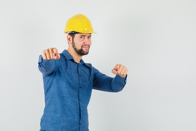 Young worker pointing at camera in shirt, helmet and looking optimistic.