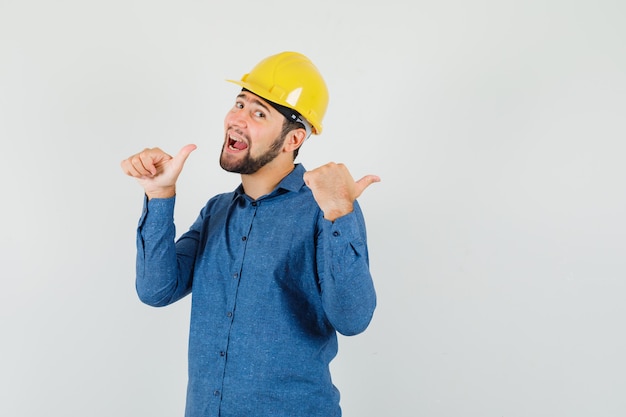 Free photo young worker pointing to the side with thumbs in shirt, helmet and looking happy