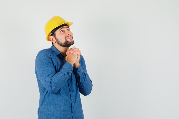 Young worker in shirt, helmet clasping hands in praying gesture and looking hopeful