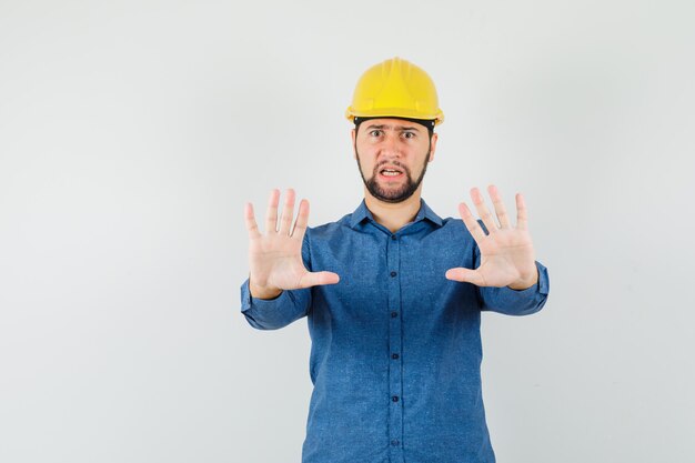 Young worker in shirt, helmet showing refusal gesture and looking irritated