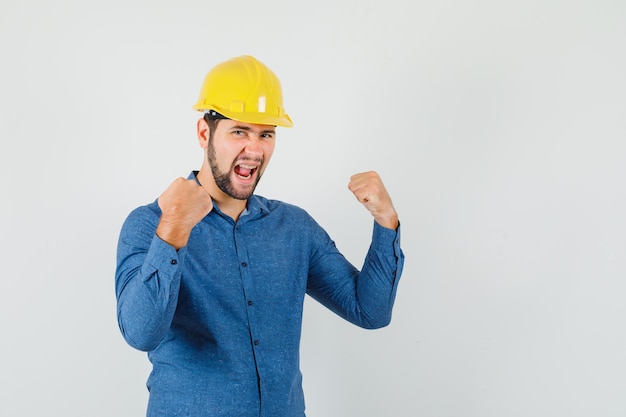 Young worker in shirt, helmet showing winner gesture and looking happy