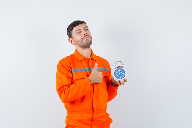 Young worker showing thumb up, holding alarm clock in uniform and looking confident.