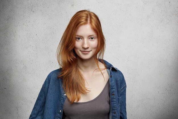 Youth and happiness. Close up view of beautiful Caucasian teenage woman with long ginger hair and freckles, dressed casually, standing against blank gray wall.