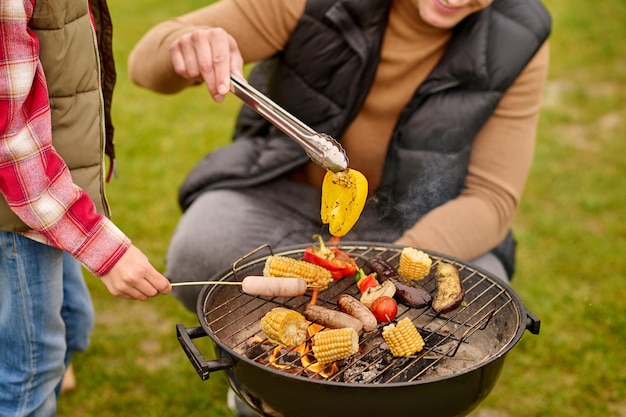 Yummy, barbecue. Man in casual clothes holding yellow pepper in tongs crouched near barbecue with vegetables and child hand with sausage on stick outdoors, no face