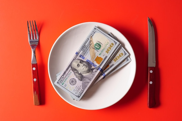 100 dollar bills on a white plate on a red background with a knife and fork