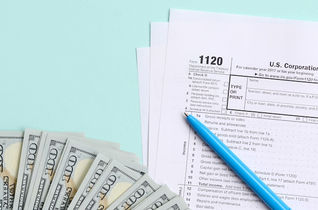 1120 tax form lies near hundred dollar bills and blue pen on a light blue