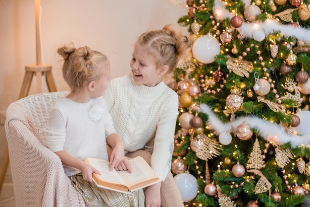 2 blonde girls are sitting on an armchair by the Christmas tree and reading a book cozy evenings with a book by the tree Girlfriends read a book together Warm relationship