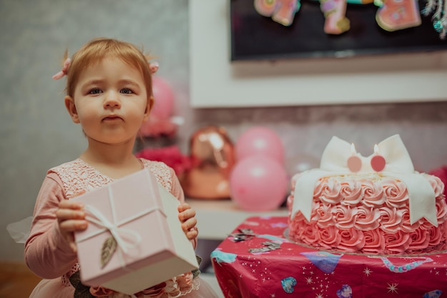 2 year baby girl in pink dress with her first birthday cake happy birthday carda cute little girl celebrates her first birthday surrounded by gifts