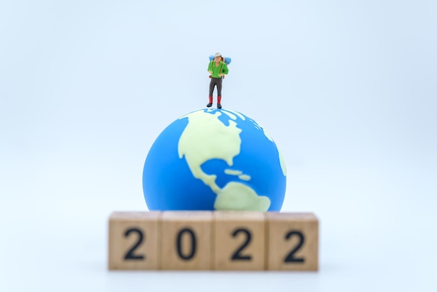 2022 New Year and Travel Concept Closeup of male traveler miniature figures with backpack standing with stack of wooden number block and mini world ball on white background