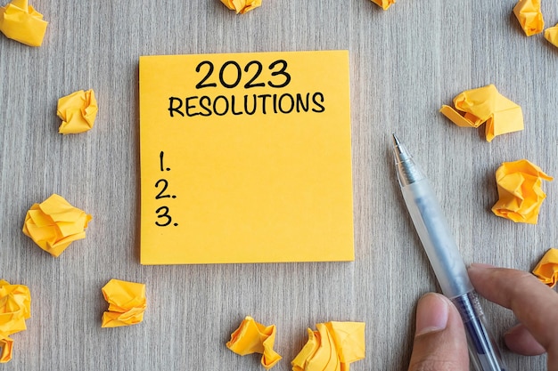 2023 RESOLUTIONS word on yellow note with Businessman holding pen and crumbled paper on wooden table background New Year start Strategy and Goal concept