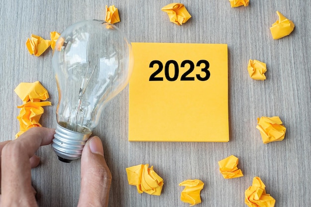 2023 words on yellow note and crumbled paper with Businessman holding lightbulb on wooden table background New Year New Idea Creative Innovation Imagination Resolution and Goal concept