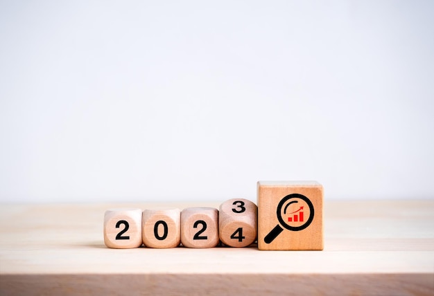 2024 happy new year with new business trends concepts Flipping the 2023 to 2024 year calendar and magnifying glass with growth graph icon on big wooden cube blocks on wood table on white background