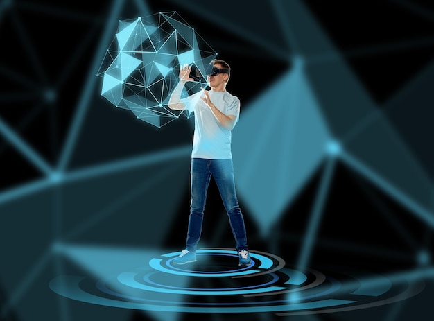 3d technology, augmented reality, gaming, cyberspace and people concept - happy young man with virtual reality headset or 3d glasses playing game and touching low poly shape hologram