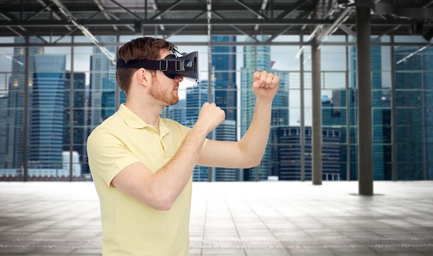 3d technology, virtual reality, entertainment and people concept - young man with virtual reality headset playing game and fighting over empty industrial room and city panorama background