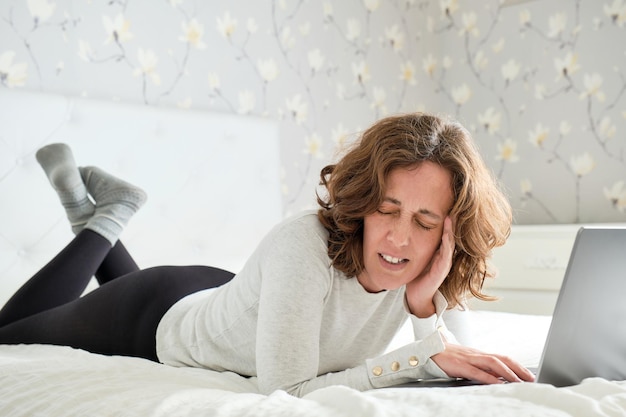 40 year old woman with a headache lying in bed trying to telework with her laptop