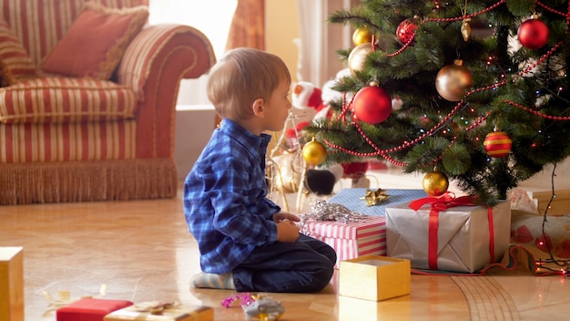 4k footage of little boy sitting on floor next to Christmas tree and looking on glowing Christmas lights and baubles. Family giving and receiving presents on winter holidays and celebrations.