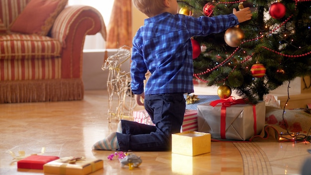 4k footage of little boy sitting on floor at living room and decorating Christmas tree with baubles and balls. Family decorating house on winter holidays and celebrations.