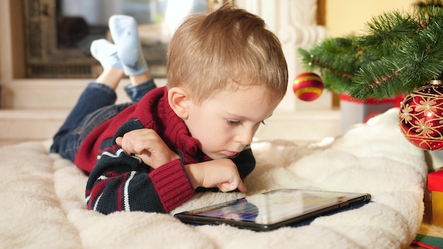 4k footage of little boy using and playing on digital computer while lying next to hristmas tree at house. Child having fun on winter holidays and celebrations.