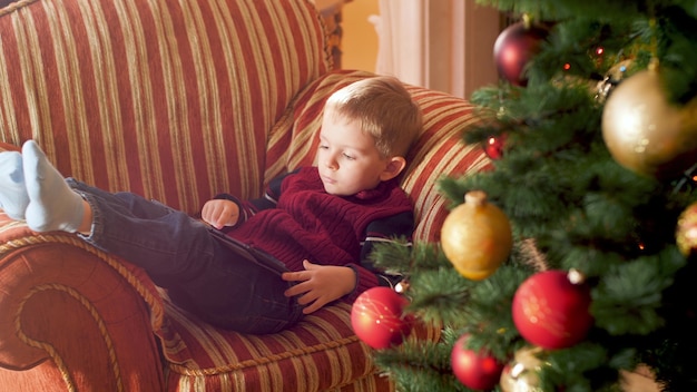 4k footage of little boy using tablet computer and browsing internet while lying on sofa next to Christmas tree. Child having fun on winter holidays and celebrations.