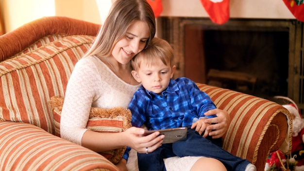 4k footage of little boy with his mother relaxing in big armchair and watching cartoons on smartphone. Family having good time and fun on winter holidays and celebrations.