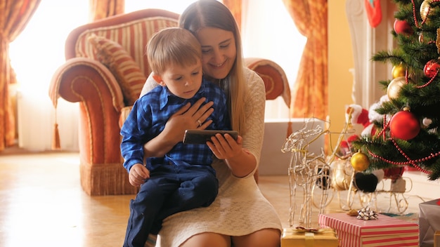 4k footage of little boy with mother watching cartoons on smartphone at Christmas morning in living room. Family having good time and fun on winter holidays and celebrations.