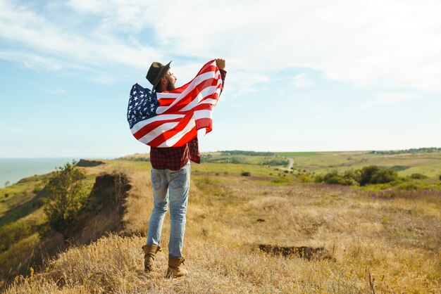 4th of July. Fourth of July. American with the national flag. American Flag. Independence Day. Patriotic holiday. The man is wearing a hat, a backpack, a shirt and jeans. 