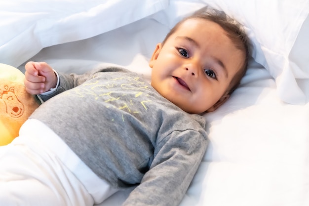 5-month-old Caucasian baby smiling in white bed just woken up, gray t-shirt and a ball to play