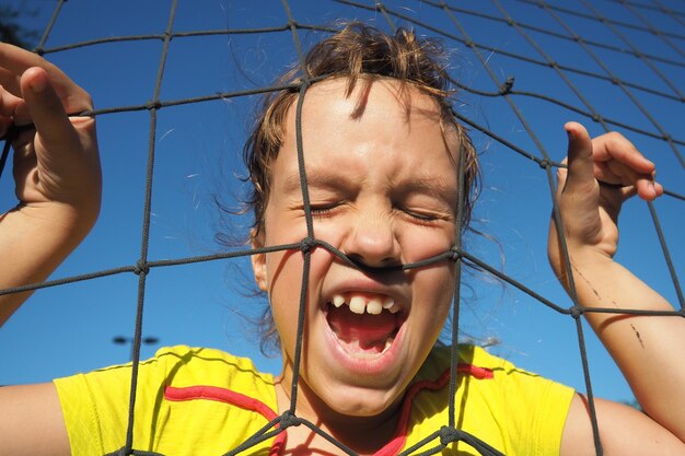 An 8yearold Caucasian girl stands behind a sports net and screams Volleyball sports dividing net against the blue sky closeup Outdoor sports