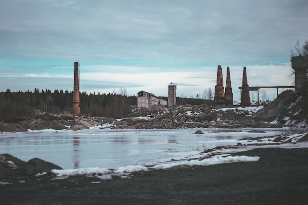 Abandoned destroyed factory in front of forest frozen river covered with ice and frost in winter