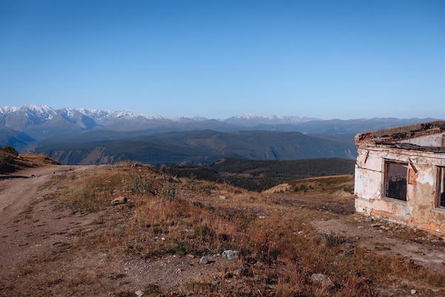 An abandoned empty small house stands on a grassy lawn near the rocky mountains under a sunny blue sky