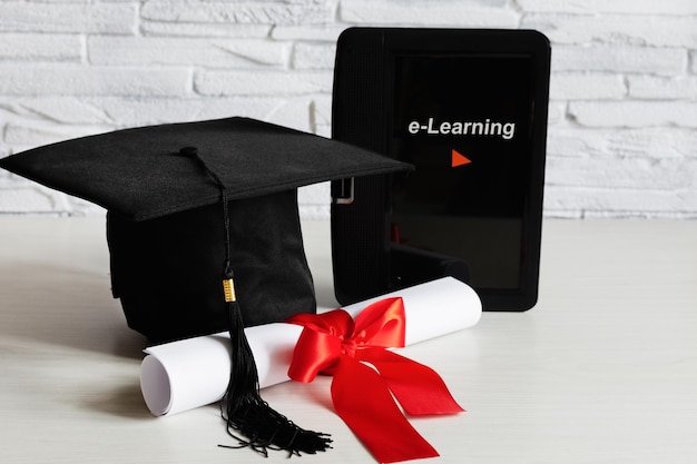 Academic cap and graduation diploma scroll and tied with red ribbon and tablet Elearning concept