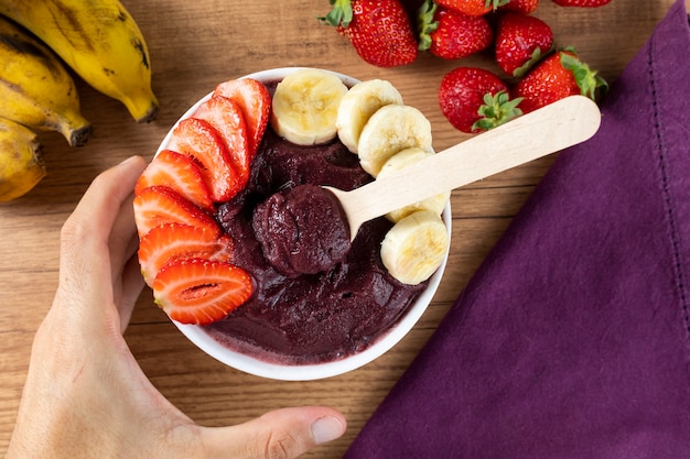 Acai, man holding an Brazilian frozen Acai berry ice cream bowl. with fruits on wooden table. Summer menu front view