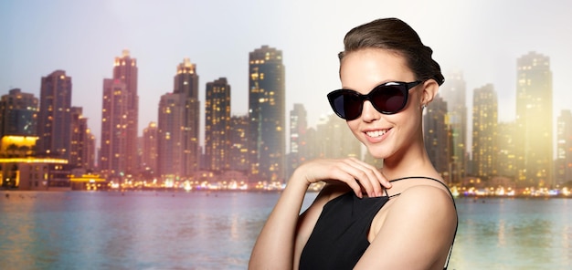 accessories, eyewear, fashion, people and luxury concept - beautiful young woman in elegant black sunglasses over night dubai city lights background