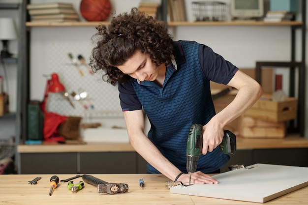 Accurate guy with dark wavy hair using electric drill for fixing metallic hinge