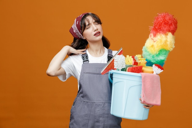 Aching young female cleaner wearing uniform and bandana holding bucket of cleaning tools keeping hand on shoulder with closed eyes isolated on orange background