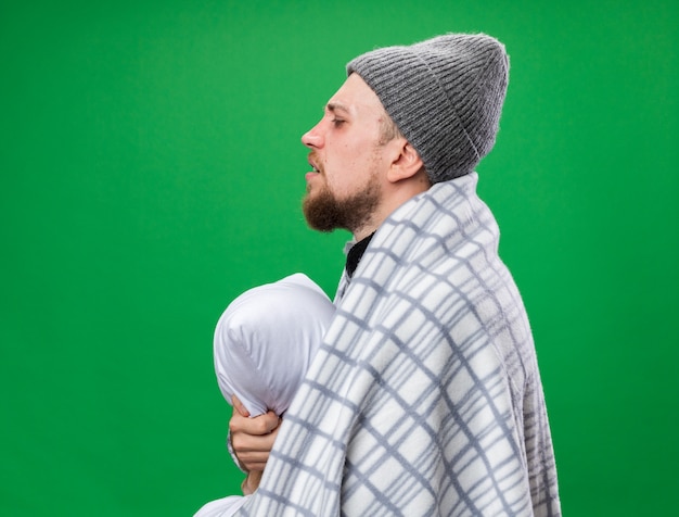 Aching young ill slavic man with scarf around neck wrapped in plaid wearing winter hat stands sideways holding pillow isolated on green wall with copy space