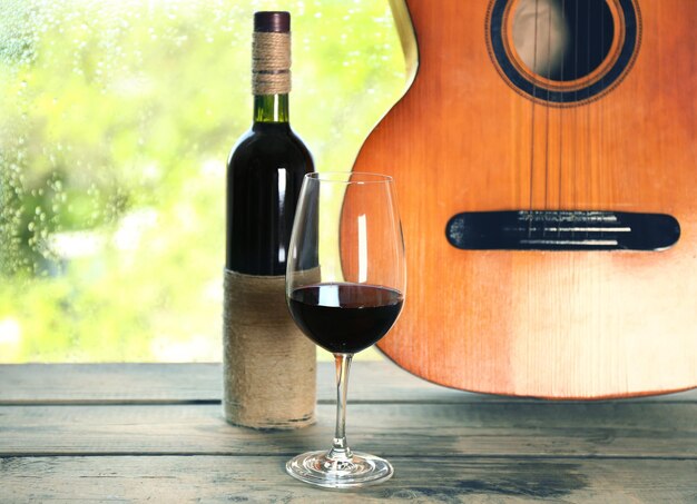 Photo acoustic guitar and glass of wine next the window with rain drops