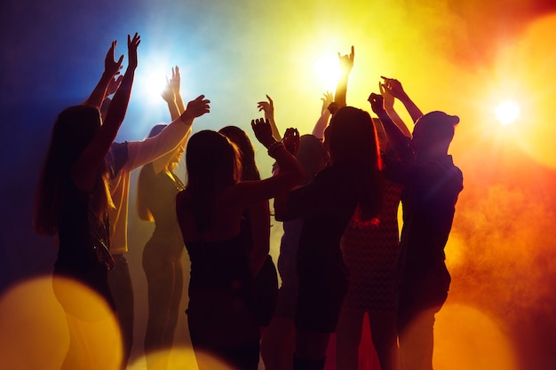 Action. A crowd of people in silhouette raises their hands on dancefloor on neon light background. Night life, club, music, dance, motion, youth. Yellow-blue colors and moving girls and boys.