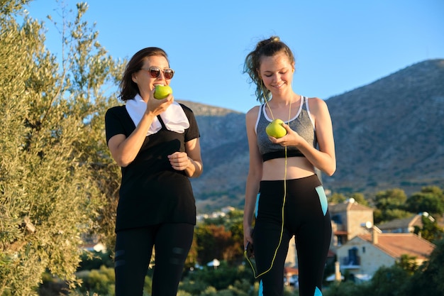 Active athletic healthy family lifestyle, smiling happy mother and teenage daughter eating apples, resting after jogging exercises. Beautiful sunset mountain nature background