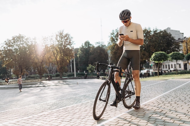 Active caucasian man in sport clothes sitting on black bike and using modern smartphone Male person using internet while taking break during workout