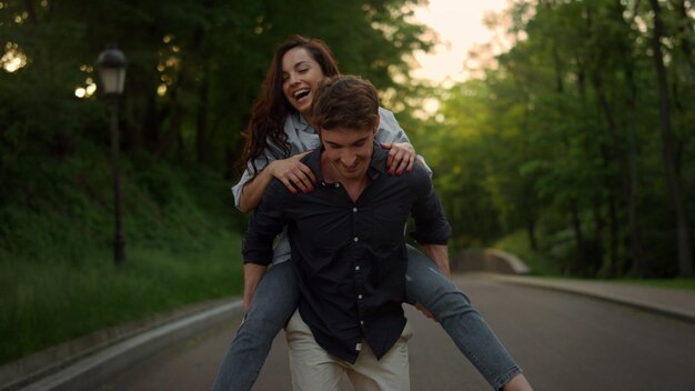 Active couple having fun during walk in summer park Handsome man carrying woman on back outdoors Young man piggyback woman on romantic date at sunset