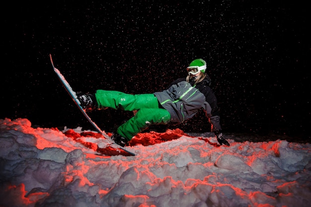 Active female snowboarder dressed in a green sportswear riding on the mountain hill in the night under the red light