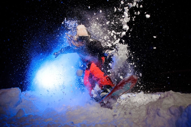 Active female snowboarder dressed in a orange sportswear jumping on the mountain slope in the night under the blue light