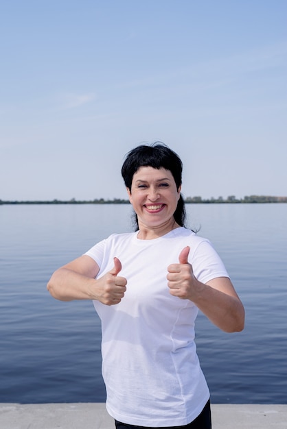 Active and happy senior woman in sportswear showing thumbs up working out near the riverside